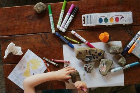 A child's hands are shown painting a rock. There are art supplies like markers, paints and paintbrushes on the table surrounding the hands.