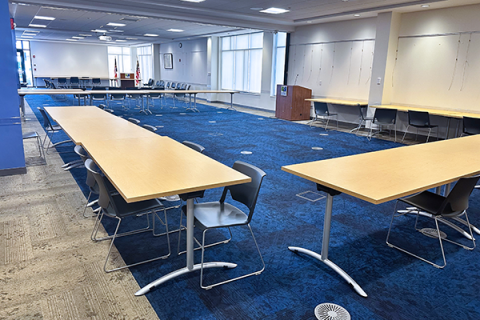 A couple of tables sit in the forefront of this image with the rest of the large, mostly empty room in the background. The partition wall is down and the image is from the back corner of the room looking towards the front. The image show clearly how large the room is.