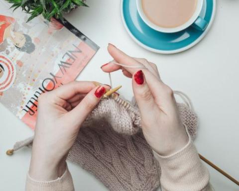 A woman's hands are shown with her fingernails painted a bright, red. She is knitting a beige scarf. There is a magazine and a cup of milky tea on the table above her hands
