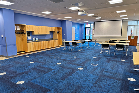 Large meeting room shown with 6 rectangular tables and 2 chairs at each table at the front of the room. The projector screen and podium are also visible at the front of the room. There is a lot of empty space in the forefront of the image to show the size of the room. To the left of the image, the door to room which leads to the main floor of the library and the cabinets and sinks are visible. 