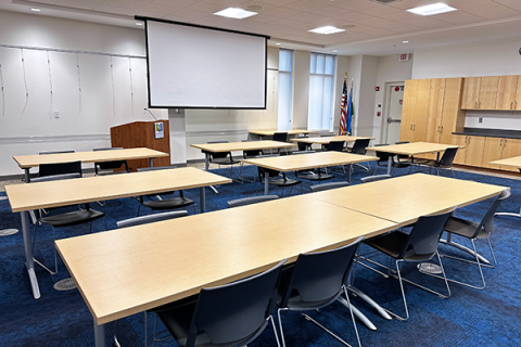 8 tables are shown with 2 chairs at each table facing the projector screen and podium.