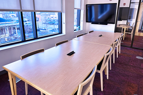 A rectangular room showing a rectangular table with 8 chairs. The table has two power adapters for charging devices built into the top. The room also has a smart board and a row of windows to the outside. The room opens onto the 2nd floor of the library by the public computers. 