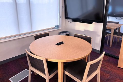 A small room with a circular table and four chairs. The table has a built-in power adapter for charging devices built into the top. There is a smart board and windows to the outside. The room opens onto the 2nd floor of the library by the public computers.