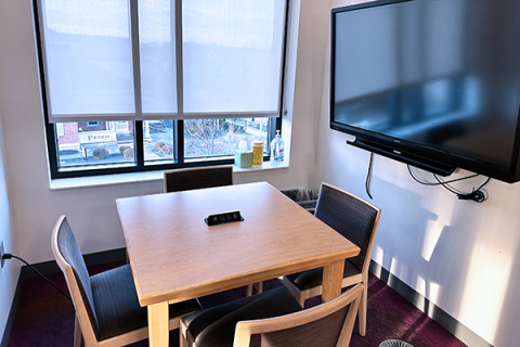 A small room with a square table and four chairs. The table has a built-in power adapter for charging devices built into the top. There is a smart board and windows to the outside. The room opens onto the 2nd floor of the library by the public computers.