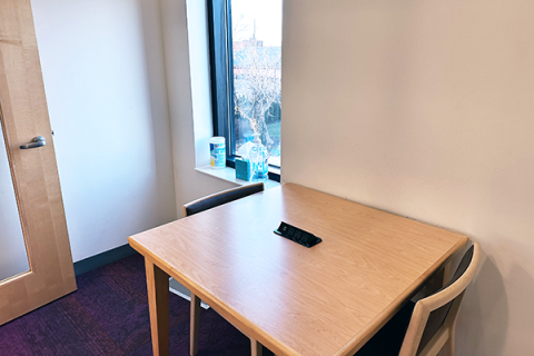 A small room with a square table and two chairs. The table has a built-in power adapter for charging devices built into the top. There is a window to the outside. The room opens onto the 2nd floor of the library by the public computers.