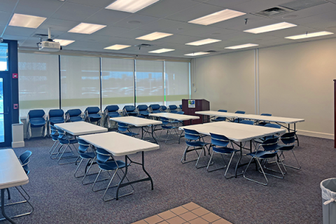 Image of Westland's meeting room. There are 8 rectangular tables up with 2 chairs at some of the tables. The podium is visible and extra chairs are stacked along the far-left bank of windows.