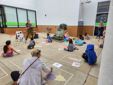 Group of children and adults sitting on sidewalk doing chalk art while receiving directions from instructor