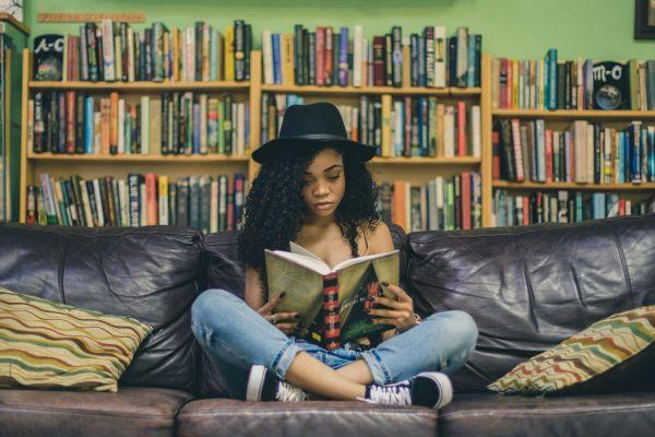 A young woman sits on a couch with her legs crossed. She is surrounded by book shelves and reading a book