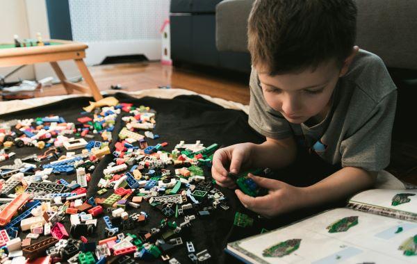 A young boys lies on his stomach in front of a bunch of LEGOs and an instruction book. He is creating something out of the LEGOs