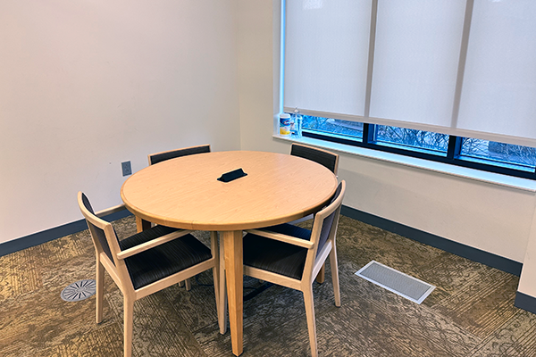 A small enclosed room showing a round table big with 4 padded chairs around it. The room has a window, and the table has a power adapter built into it.