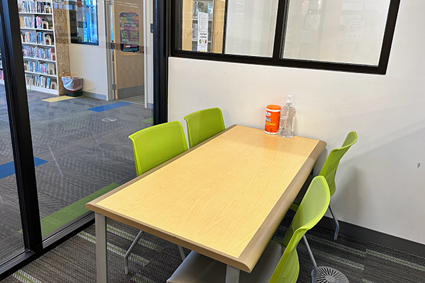 A small, interior room with a rectangular table and 4 green chairs. The room is surrounded by windows opening up to the teen room and A/V area of the library.