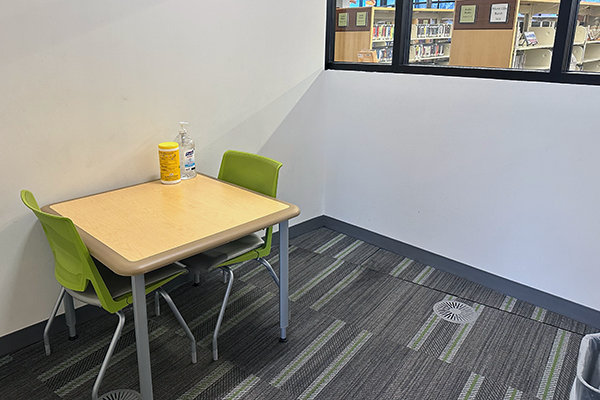 A small, interior room with a small, square table and two green chairs. The room opens into the teen area and has a window to the A/V area of the library.