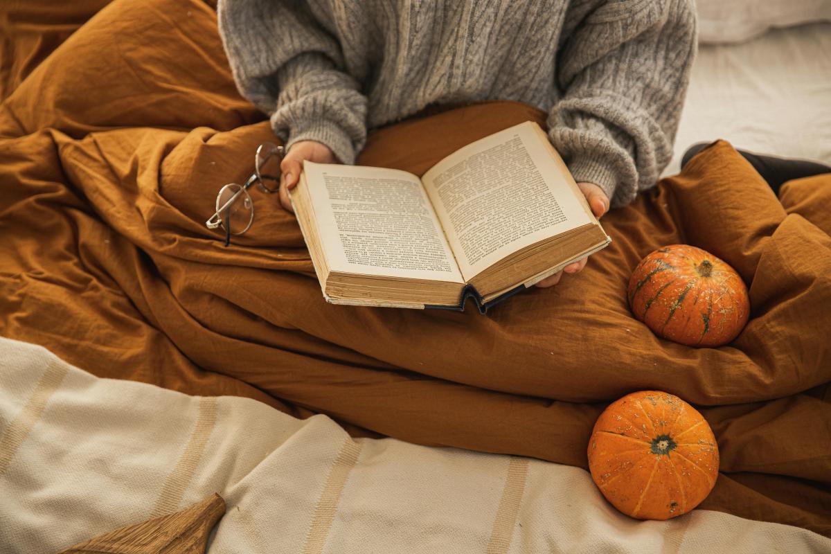 fall-themed photo of someone reading a book while seated with a blanket and with two mini pumpkins used as decorative props